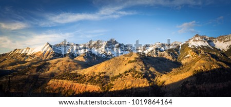 Panoramic view of a mountainside lit by a setting sun in the rocky mountains.  The glow of the sun illuminates the fall colors, along with the first snow found high up in the mountain peaks. 