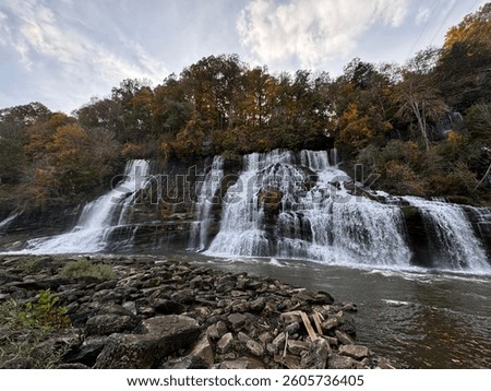 Panoramic view of a multi-tiered waterfall cascading down a rocky cliff surrounded by autumn trees, flowing into a river lined with large stones. Dramatic natural landscape under a moody evening sky 