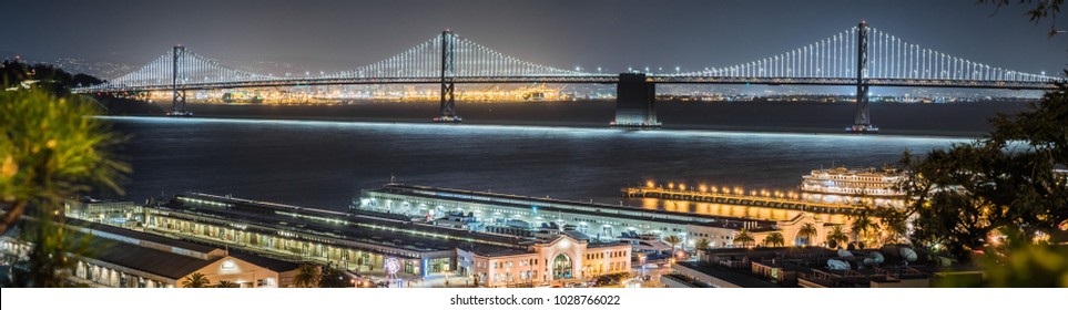 Panoramic View Of The Night Illuminated Bay Bridge Connecting San Francisco And Oakland; Ferry Terminals And Ptehr Piers In The Foreground; Night Photography; San Francisco Bay Area, California