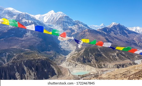 A panoramic view on Manang valley from Praken Gompa, Nepal. Prayer flags waving above the snow capped peaks of Annapurna Chain. There is a small lake in the valley. Harsh landscape. Serenity