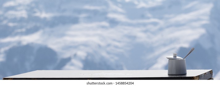 Panoramic View On Old Table With Sugar Bowl In Outdoor Cafe At Ski Resort And Snowy Mountains At Background