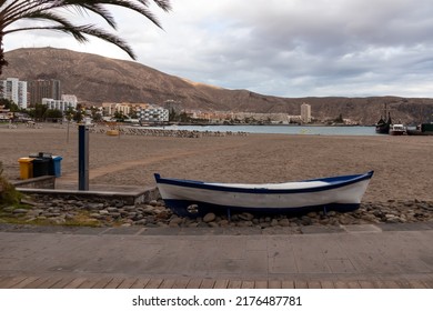 Panoramic View On Sand Beach Of Playa De Los Cristianos On Tenerife, Canary Islands, Spain. A Decorative Wooden Boat On The Side Of The Touristic Promenade Street. Small Barren Hills And City View