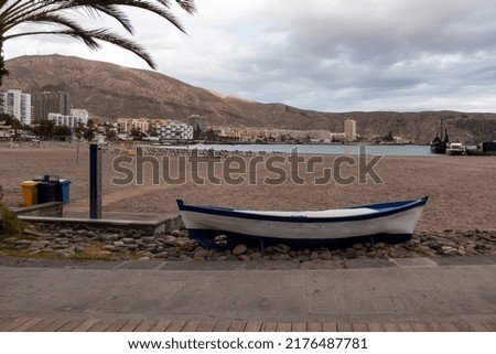 Panoramic view on sand beach of Playa de Los Cristianos on Tenerife, Canary Islands, Spain. A decorative wooden boat on the side of the touristic promenade street. Small barren hills and city view