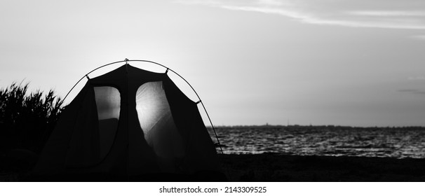 Panoramic View On Sea Coast And Camping Tent With Shining Through Entrance On Beach At Summer Evening. Black And White Toned Landscape. High Contrast.