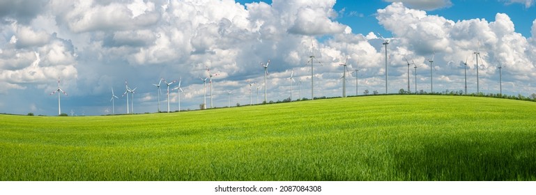 Panoramic View Over Beautiful Farm Landscape With Wheat Field As A Wave And Wind Turbines To Produce Green Energy In Germany, Summer, At Sunny Day And Blue Cloudy Sky