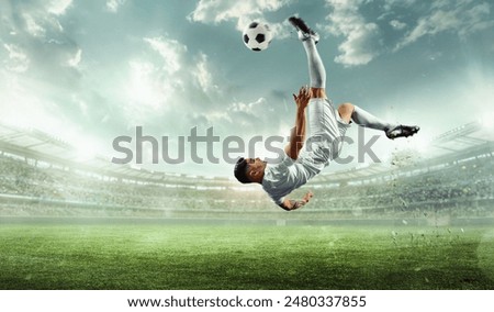 Panoramic view of performs impressive kick, caught mid-air with ball high above him against stadium illuminated by spotlights. Concept of sports games, competition, championship, tournament. Ad