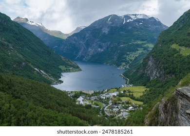 A panoramic view of a picturesque Norwegian fjord, showcasing towering mountains, lush greenery, a winding river, and a small village nestled at the foot of the mountains. Geiranger fjord Norway - Powered by Shutterstock