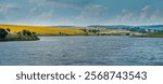 A panoramic view of a reservoir, hills with fields of sunflowers and lush greenery under a bright blue sky with clouds. Quiet countryside by a lake or river in summer.