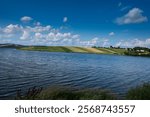 A panoramic view of the reservoir, hills in lines with fields of green grass, under a bright blue sky with clouds. Quiet countryside by a lake or river in summer.