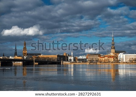 Panoramic view of Riga, Latvia, featuring the historic skyline reflected in the calm waters of the Daugava River beneath dramatic spring clouds.