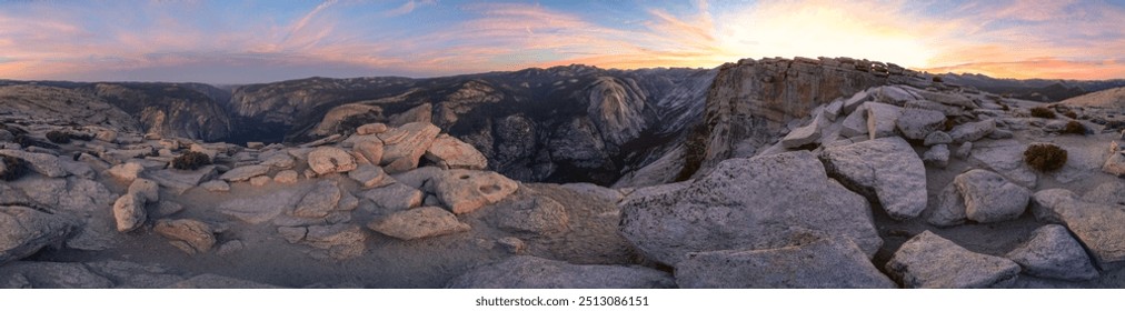 A panoramic view of a rocky mountain range with a beautiful sunset in the background. The sky is filled with clouds, and the sun is setting, casting a warm glow over the landscape