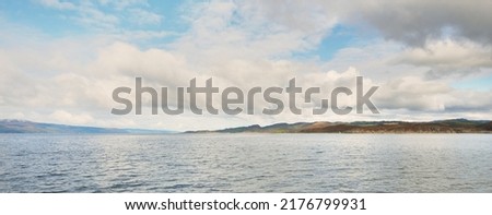 Panoramic view of the rocky river shores from the water. Trees, hills and mountains in the background. Cloudy blue sky. Gare Loch, Firth of Clyde, Scotland, UK