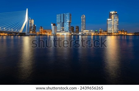 Panoramic view of Rotterdam night time skyline with modern buildings and iconic bridge. Evening sky reflection in River Maas at blue hour with colorful illumination. Major port and big dutch metropole