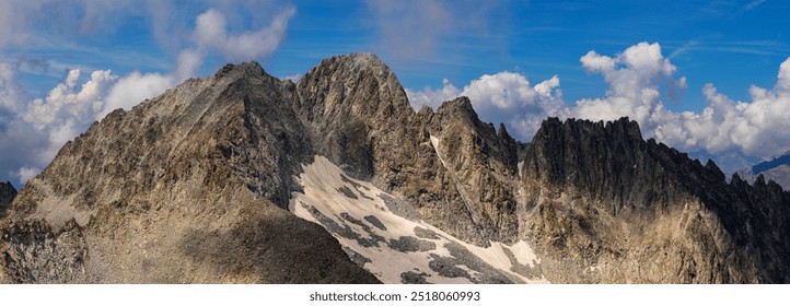A panoramic view of a rugged mountain range partially covered by clouds, with rocky peaks and alpine terrain extending into the distance.