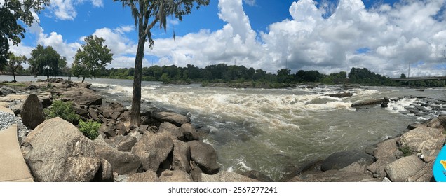 A panoramic view of a rushing river with rapids surrounded by large rocks and lush greenery under a vibrant blue sky with scattered white clouds. 