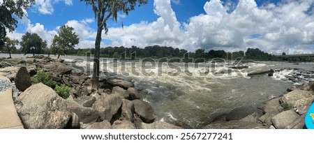 A panoramic view of a rushing river with rapids surrounded by large rocks and lush greenery under a vibrant blue sky with scattered white clouds. 