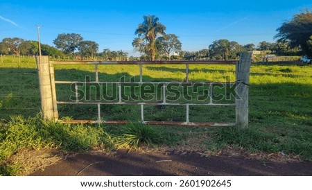 A panoramic view of a rustic metal gate with a lush green pasture in the background. The landscape features tall trees, a clear blue sky, and soft shadows cast across the path.