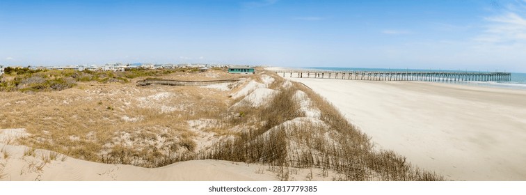 A panoramic view of a sandy beach with dunes and coastal vegetation leading to a wooden boardwalk and a long pier extending into the Atlantic Ocean under a clear blue sky