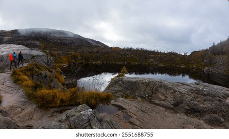 A panoramic view of a serene lake surrounded by rocky terrain and autumn foliage. Two hikers are seen walking along the rocky edge, with misty mountains in the background under a cloudy sky. - Powered by Shutterstock