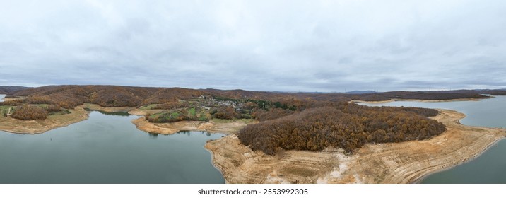 Panoramic view of a serene lake surrounded by autumnal forests.An aerial shot of a lake with a shoreline of dried, light brownish-tan earth, surrounded by dense forests displaying the colors of autumn - Powered by Shutterstock