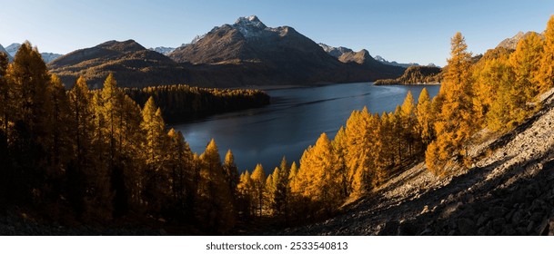 A panoramic view of a serene mountain lake surrounded by autumn trees and snow-capped peaks under a clear blue sky. - Powered by Shutterstock