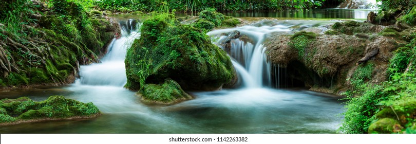 Panoramic view of small waterfalls streaming into small pond in green forest in long exposure - Shutterstock ID 1142263382