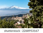 Panoramic view of snow capped Mount Etna volcano on a sunny day seen from Taormina, Sicily, Italy, Europe, EU. Tree and cactus on the side. Overview of the hilltop city of Taormina. Mountain landscape