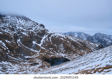 Panoramic view of a snow-capped mountain range with a frozen lake in the valley. The sky is overcast and the air is crisp. Făgăraș Mountains, Romania - Powered by Shutterstock