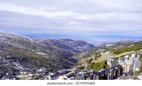 A panoramic view of a snow-capped mountain range with a small ski resort nestled in a valley. The sky is cloudy, but the scenery is still breathtaking.