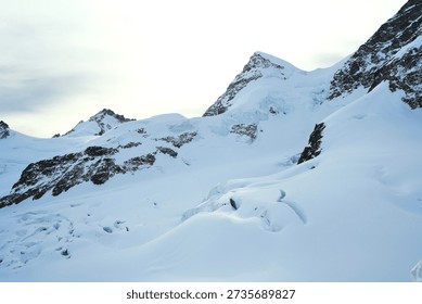 Panoramic view of a snow-capped mountain range under a soft, overcast sky. The rugged, rocky peaks are heavily draped in white snow and blue-tinted glacial ice
