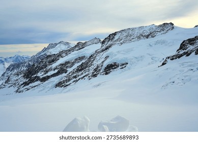 Panoramic view of a snow-capped mountain range under a soft, overcast sky. The rugged, rocky peaks are heavily draped in white snow and blue-tinted glacial ice