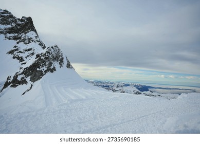 Panoramic view of a snow-capped mountain range under a soft, overcast sky. The rugged, rocky peaks are heavily draped in white snow and blue-tinted glacial ice