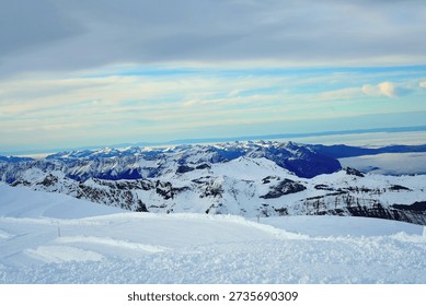 Panoramic view of a snow-capped mountain range under a soft, overcast sky. The rugged, rocky peaks are heavily draped in white snow and blue-tinted glacial ice