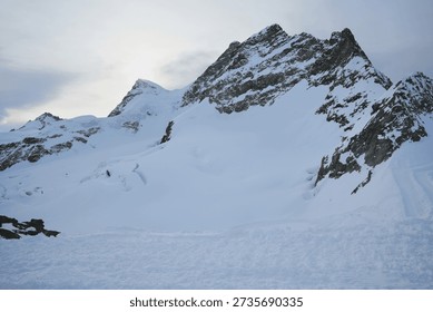 Panoramic view of a snow-capped mountain range under a soft, overcast sky. The rugged, rocky peaks are heavily draped in white snow and blue-tinted glacial ice