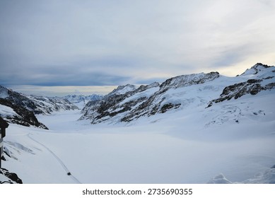 Panoramic view of a snow-capped mountain range under a soft, overcast sky. The rugged, rocky peaks are heavily draped in white snow and blue-tinted glacial ice