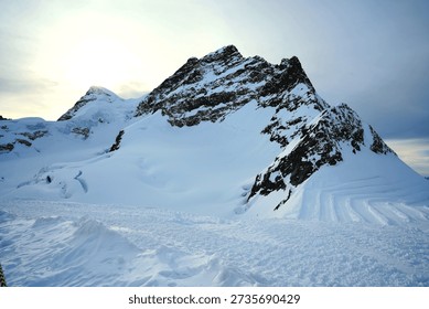 Panoramic view of a snow-capped mountain range under a soft, overcast sky. The rugged, rocky peaks are heavily draped in white snow and blue-tinted glacial ice