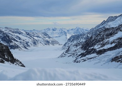 Panoramic view of a snow-capped mountain range under a soft, overcast sky. The rugged, rocky peaks are heavily draped in white snow and blue-tinted glacial ice