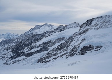 Panoramic view of a snow-capped mountain range under a soft, overcast sky. The rugged, rocky peaks are heavily draped in white snow and blue-tinted glacial ice