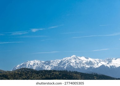 Panoramic view of snow-covered mountain range above forested hills under a clear blue sky. Bright daylight landscape with wide copy space, ideal for travel, nature, and outdoor themes.