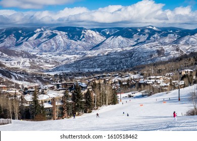 Panoramic view of Snowmass Village, with skiers skiing at the Aspen Snowmass ski resort in the foreground and the Rocky Mountains of Colorado in the background, on a partly cloudy winter day.  