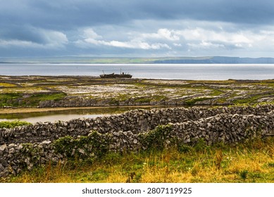 Panoramic view of stone walled fields leading to a rocky shoreline with a rusted shipwreck on the coast of Inisheer in the Aran Islands, Ireland. The Atlantic Ocean and distant cliffs are visible under a dramatic cloudy sky, with green grass and limestone rocks in the foreground creating a textured rural landscape.