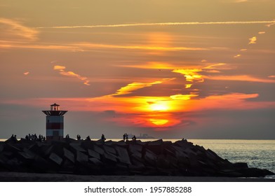 Panoramic view of sun setting in North Sea from the beach of Scheveningen with in forefront the Northern Pier with iconic red-white striped harbor light and silhouette of people enjoying the sunset