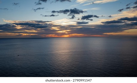 A panoramic view of the sun setting on the horizon over the calm sea waters. A small boat leaves a solitary trail on the silvery surface, while the sky is painted in shades of orange, yellow, and blue with scattered clouds. An evocative image capturing the peace, solitude, and natural beauty of the Sardinian coast.