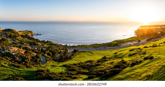 Panoramic view of the sun setting over the Pacific Ocean, casting a warm glow on the coastal cliffs and lush green landscape of Rancho Palos Verdes
