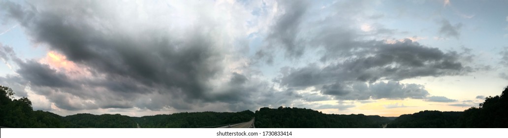 Panoramic view of sunset colors with clouds over empty road with trees and hills around it 