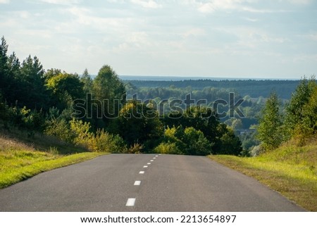 Panoramic view from the top of the hill to the road and the autumn forest on the horizon. An empty asphalt road runs through a hilly area through a forest and a village. Empty space for text