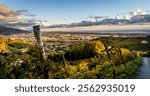 A panoramic view of a valley with vineyards, rolling hills, and a coastal town in the distance under a cloudy sky during sunset.