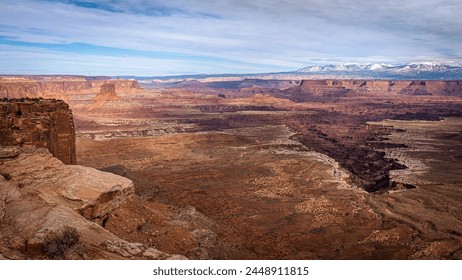 Panoramic view of a vast canyon with layered red rock formations under a partly cloudy sky. Snow-capped mountains can be seen in the distance, adding contrast to the arid landscape in the foreground. - Powered by Shutterstock