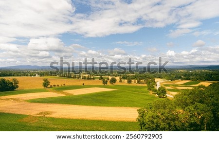 A panoramic view of a vibrant patchwork of farmland, including golden and green fields, a winding country road, and a backdrop of rolling mountains under a sunny sky.