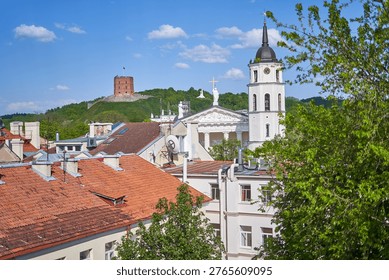 A panoramic view of Vilnius, Lithuania, showcasing the city's historic architecture and landmarks. In the foreground, traditional red terracotta rooftops of old town buildings are visible with chimneys and architectural details. The neoclassical Vilnius Cathedral with its prominent white bell tower, clock, and columns stands in the middle ground, topped with a golden cross and white statues. Behind the cathedral, the green hill of Gediminas Castle rises, featuring the iconic red-brick Gediminas Tower, a medieval fortification and remaining part of the Upper Castle. White crosses are visible on the adjacent hilltop. Lush green trees frame the scene on the right side, while a bright blue sky with scattered white clouds provides a vibrant backdrop. The image captures the blend of religious architecture, medieval fortifications, and residential buildings characteristic of the Lithuanian capital's historic district.